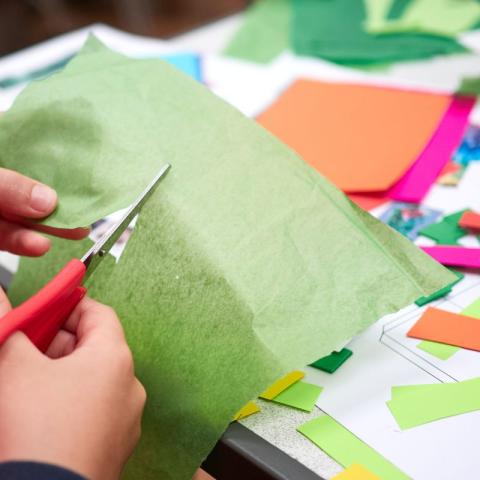 Hands use red scissors to cut green tissue paper on a table covered with craft supplies.