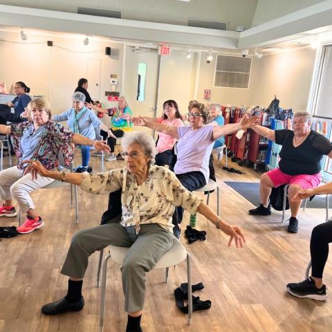 A group of older adult women participates in a seated exercise class, extending their arms out horizontally.